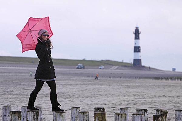 junge Frau mit rosa Schirm balanciert ueber Buhnen, Leuchtturm im Hintergrund, young woman with pink umbrella balancing on spur dikes with lighthouse in the background