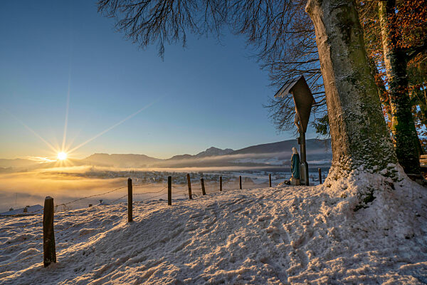 winterlicher Sonnenaufgang mit Wegkreuz über Teisendorf mit dem Untersberg, den Bergen hinter Salzburg und in der Mitte dem Hochstaufen im Hintergrund Deutschland
gesehen vom Schelmberg aus