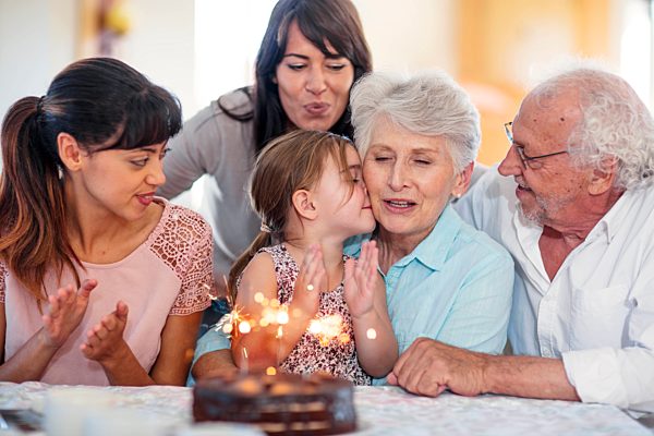 Little girl lwatching sparklers on a birthday cake, sitting on grandmother's lap, with family around