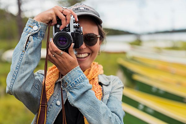 Finland, Lapland, portrait of smiling young woman taking picture with a camera at the lakeside