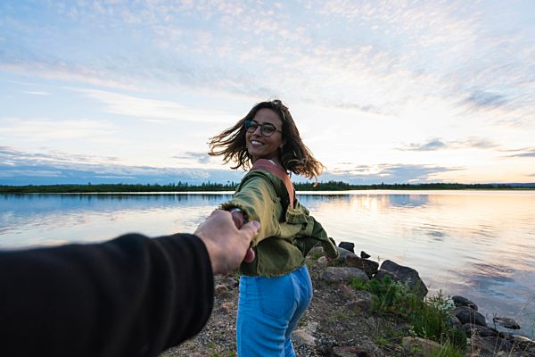 Finland, Lapland, happy young woman holding man's hand at the lakeside at twilight
