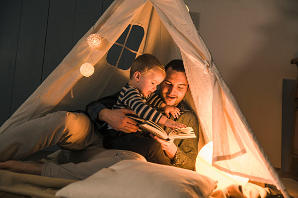 Father reading book to son at an illuminated tent at home