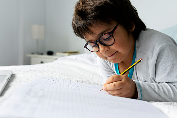 Portrait of little boy lying on bed doing homework