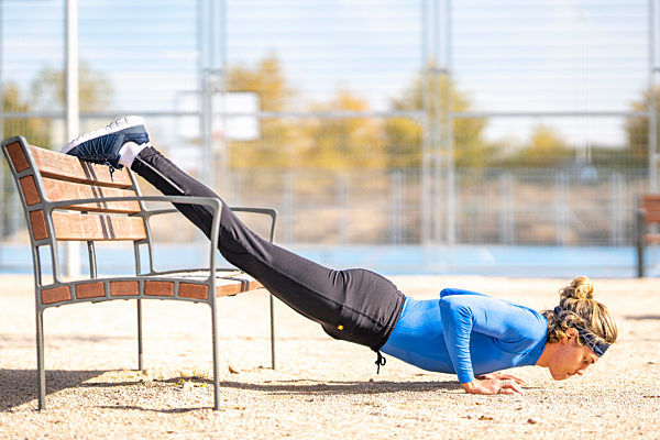 Male runner doing push ups with dedication on bench in public park on sunny day