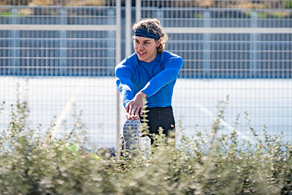 Young male sportsperson looking away while stretching in public park on sunny day