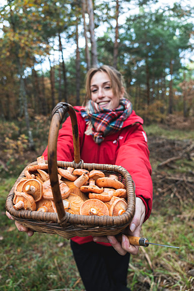 Smiling woman showing basket full of mushrooms in forest during autumn