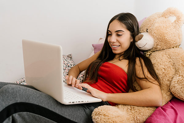 Smiling teenage girl using laptop while leaning on teddy bear toy at home
