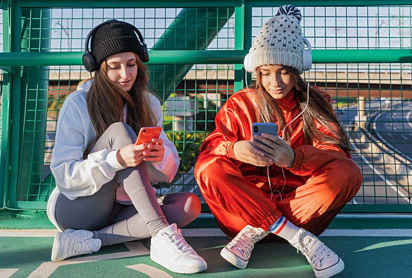 Friends wearing knit hat and and headphones using mobile phones while sitting on bridge