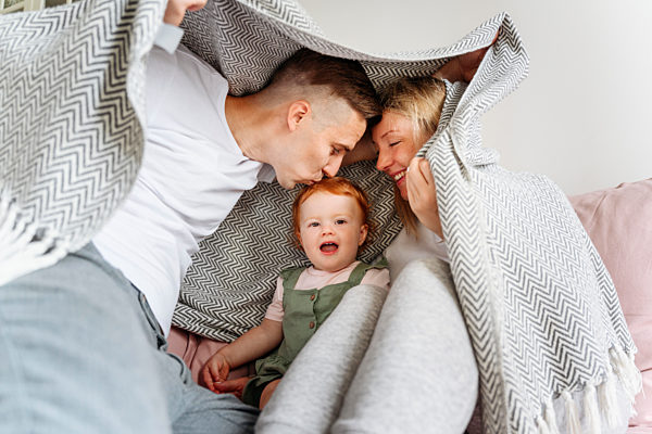 Happy family with baby daughter hiding under blanket on sofa