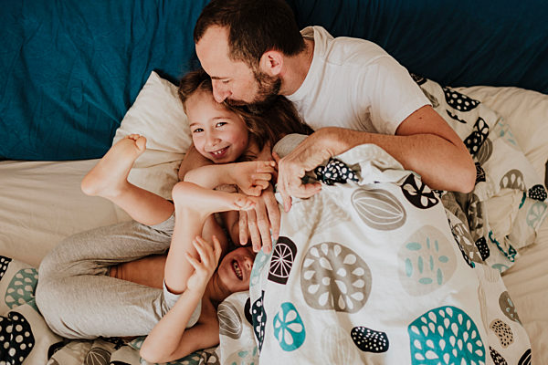 Father kissing daughter on forehead while son laughing on bed at home