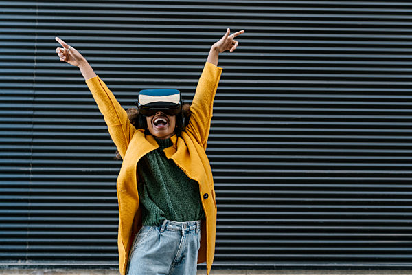 Cheerful woman with arms raised gesturing while watching video through wearable computer against wall