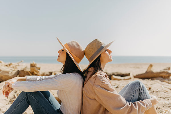 Female twins sitting back to back on sand at beach during sunny day