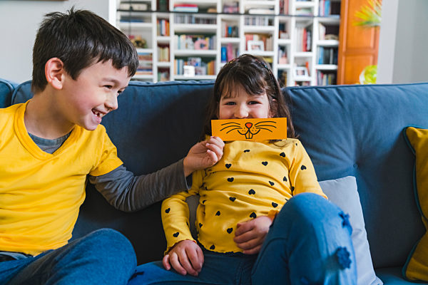 Smiling boy holding emoticon over girl's face while sitting on sofa at home