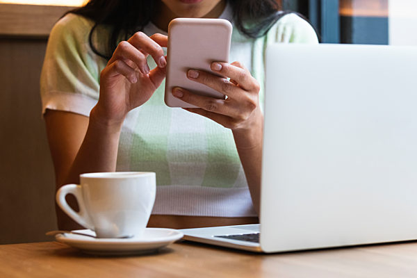 Woman with laptop and coffee cup using smart phone at cafe