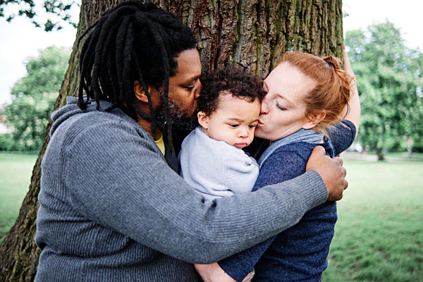 Father and mother kissing son while embracing near tree trunk at park