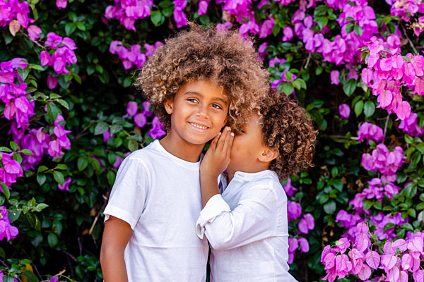 Smiling boy looking away while brother gossiping in ear in front of tree