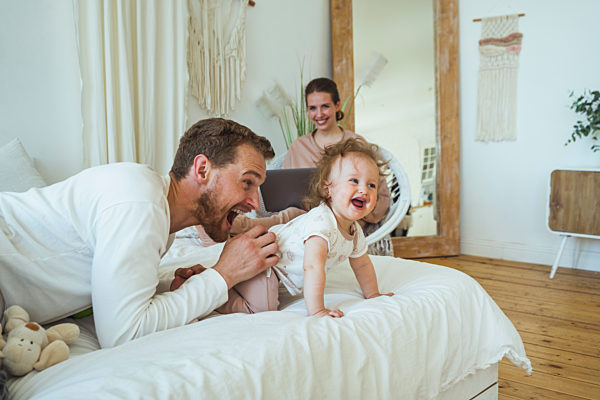 Smiling woman looking at cheerful man playing with daughter in bedroom