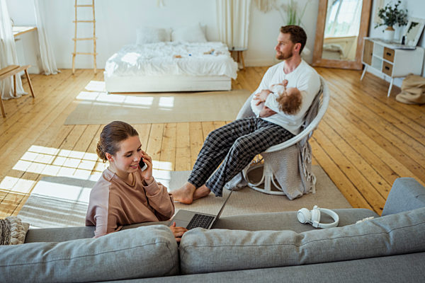 Businesswoman with laptop talking on mobile phone while man carrying daughter on chair at home