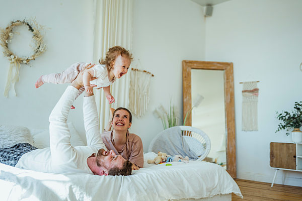 Happy woman looking at cheerful daughter picked up by man on bed at home