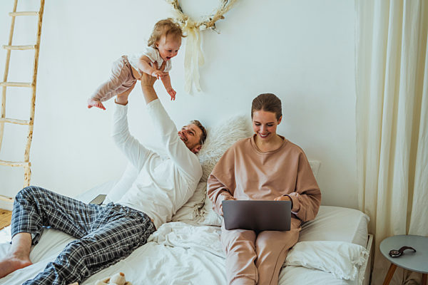 Smiling woman using laptop by playful man lifting daughter on bed at home