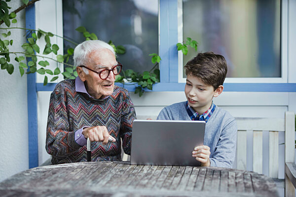 Senior man talking to grandson sitting with tablet PC at table
