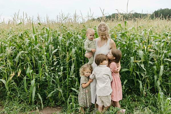 Young woman carrying toddler boy embracing daughters and son in corn field