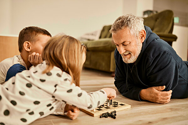 Grandfather looking at girl playing chess lying on floor at home