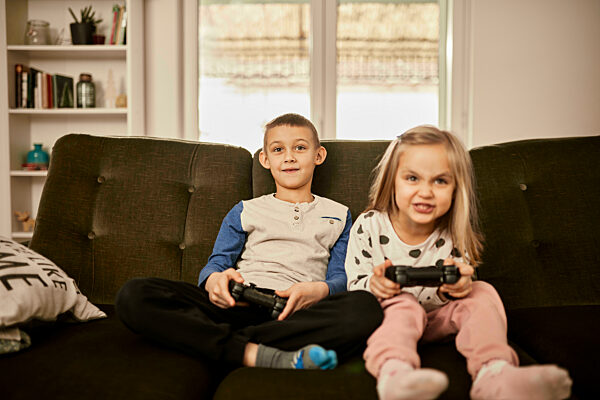 Brother and sister playing video game sitting on sofa at home