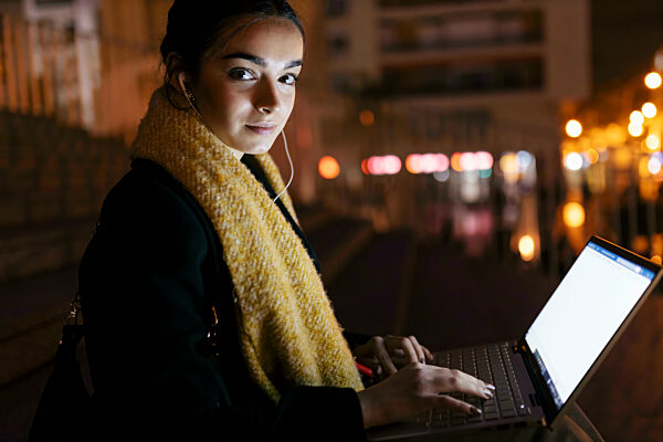 Teenage girl with laptop listening music on in-ear headphones at night