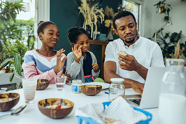 Father and daughters on video call through laptop at breakfast table