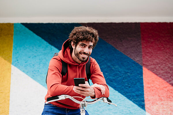Smiling man with smart phone on bicycle in front of colorful wall