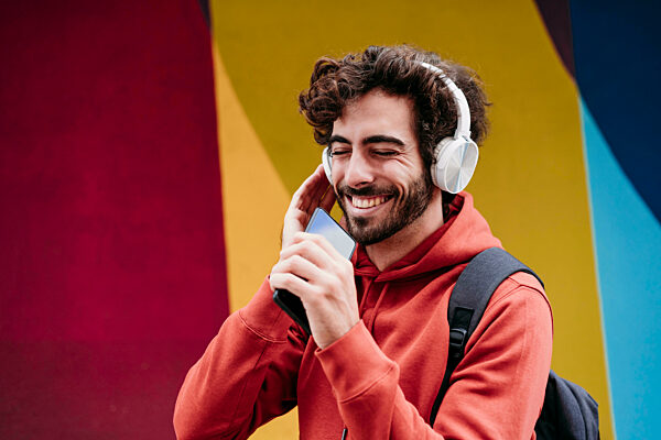 Happy man with eyes closed listening to music holding smart phone in front of colorful wall