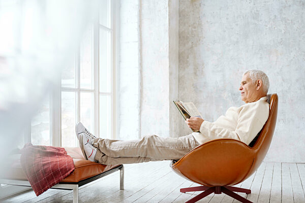 Senior man reading book sitting on chair at home