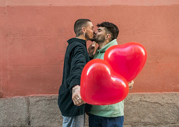 Affectionate gay couple kissing with red heart shape balloon in front of wall