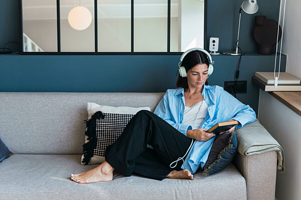 Young woman with book listening to music through headphones at home