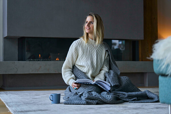 Smiling woman wrapped in blanket sitting with book at home