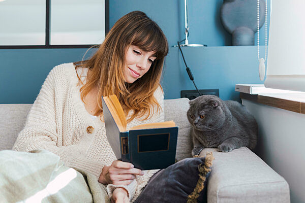Woman showing book to cat sitting on sofa at home
