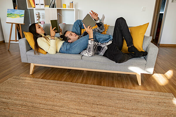 Woman reading book with boyfriend using tablet PC lying on sofa at home