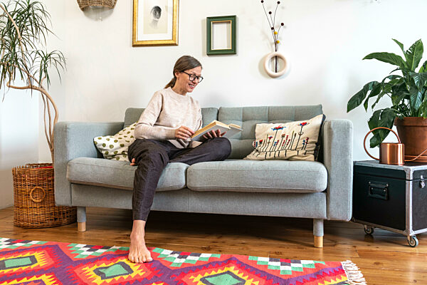 Smiling mature woman sitting on sofa and reading book at home