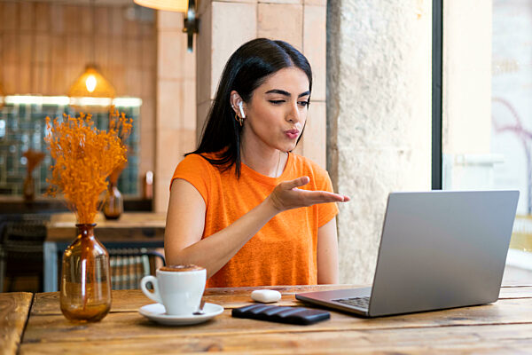 Young woman blowing kiss on video call through laptop sitting in cafe
