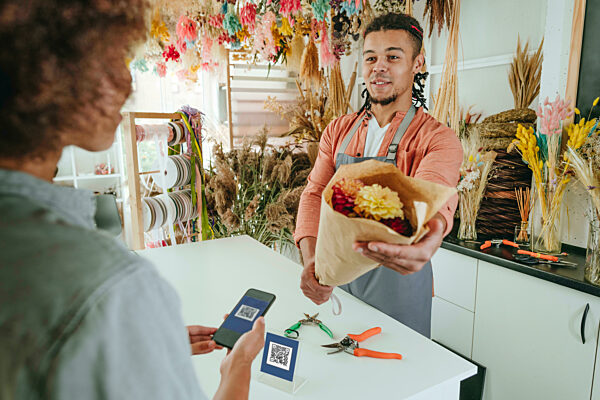 Smiling young florist giving flower bouquet to customer paying through contactless method at store