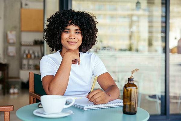 Smiling woman with curly hair writing on note pad at sidewalk cafe