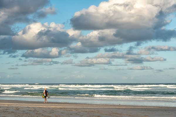 Australia, New South Wales, Pottsville, angler walking along beach with surf and dark clouds