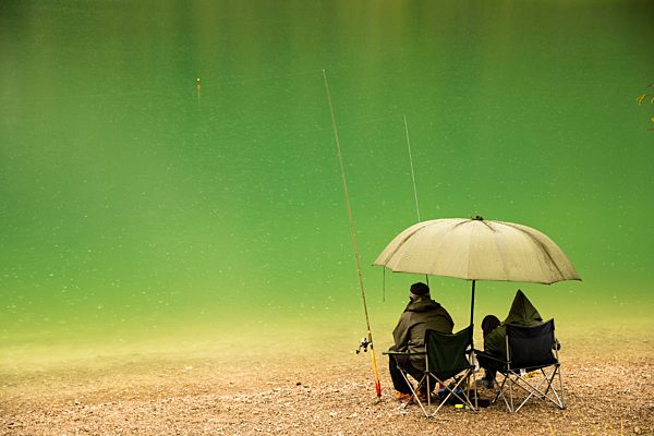 Austria, Tyrol, Lake Plansee, two anglers sitting at lakeshore in the rain