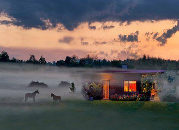 Estonia, lighted wooden house and horses in rural misty landscape