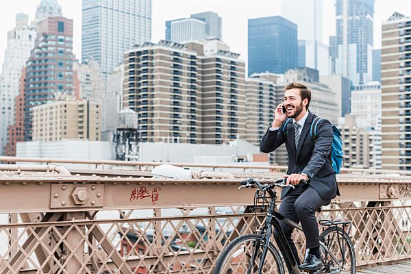 USA, New York City, laughing businessman on bicycle on Brooklyn Bridge using cell phone