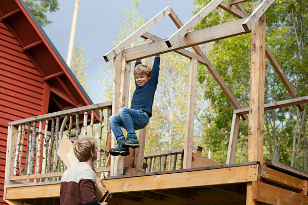 Young boy beams with joy as he plays on a wooden playset on a sunny day, with another child observing nearby.
