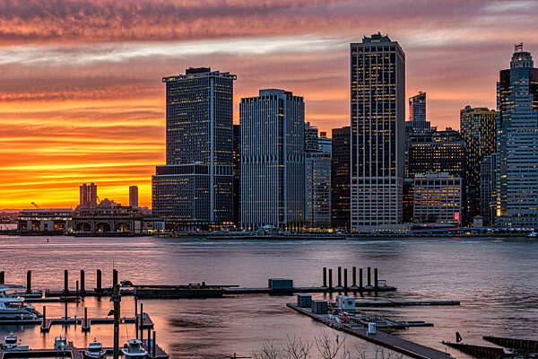 Vibrant sunset over a city skyline with tall buildings and a calm river in the foreground. New York City, NY, USA