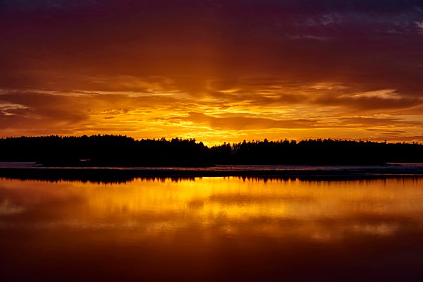 Dramatic Glowing Cloud Reflected In The Tranquil Ocean Water At Sunset With A Silhouette Of Trees Along The Coastline At Mackenzie Beach, Vancouver Island; Tofino, British Columbia, Canada