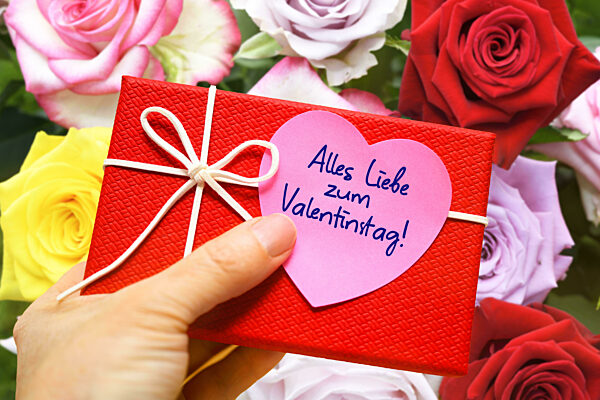 Hand Holding Gift With A Heart Shaped Note And Inscription All Love For Valentines Day In Front Of A Bouquet Of Flowers